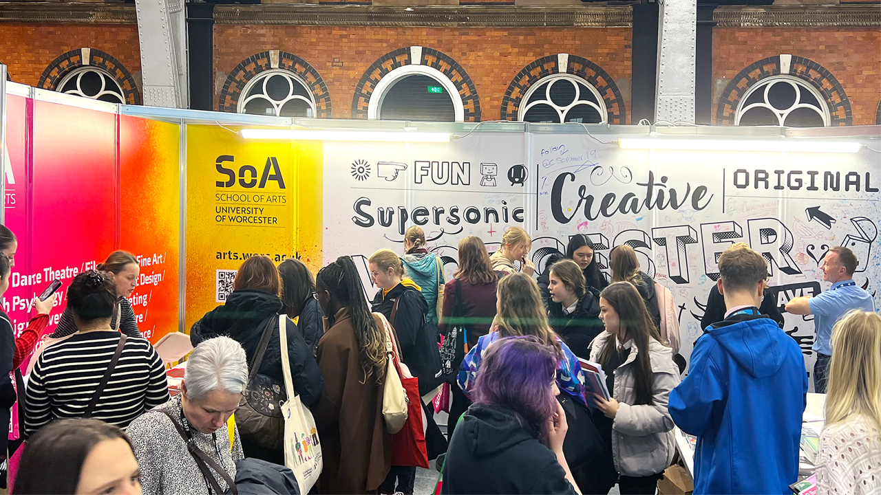 Image of University of Worcester's stand at UCAS. The long back wall has adjectives in varying fonts, and blends into a yellow, orange, and pink splattered side wall covered with names of course titles that the University offers. The stand is very busy with many people around, and the back wall is covered in drawings that visitors have contributed. 